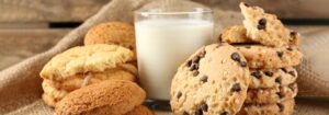 Vaso de leche fresca rodeado de galletas con chispas de chocolate y galletas de avena sobre un mantel de yute.