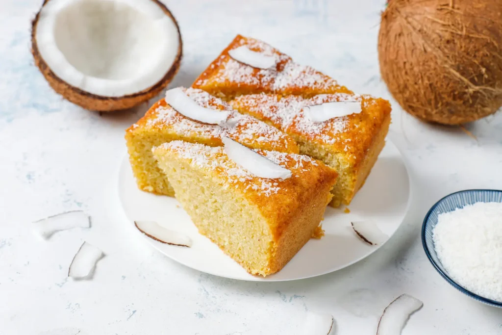 Porciones de pastel de coco espolvoreadas con coco rallado, junto a un coco entero y partido sobre un fondo claro.