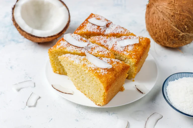 Porciones de pastel de coco espolvoreadas con coco rallado, junto a un coco entero y partido sobre un fondo claro.