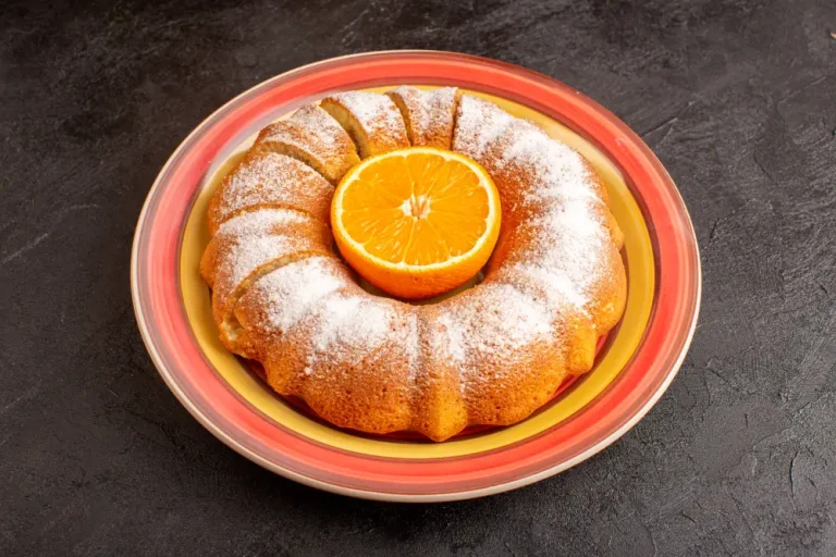 Torta de naranja tipo Bundt espolvoreado con azúcar glas en un plato decorado, con media naranja fresca colocada en el centro.